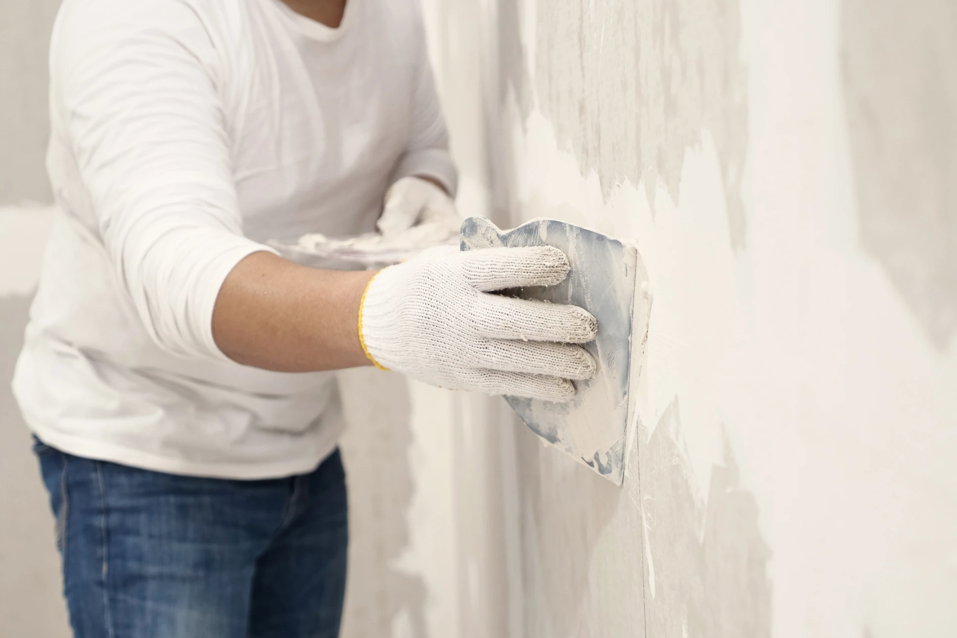 Main tirant un enduit de lissage sur un mur blanc avec une spatule métallique