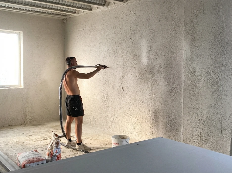 Ouvrier torse nu applique un enduit de projection mécanisé sur mur intérieur brut, tenant un flexible de machine à projeter